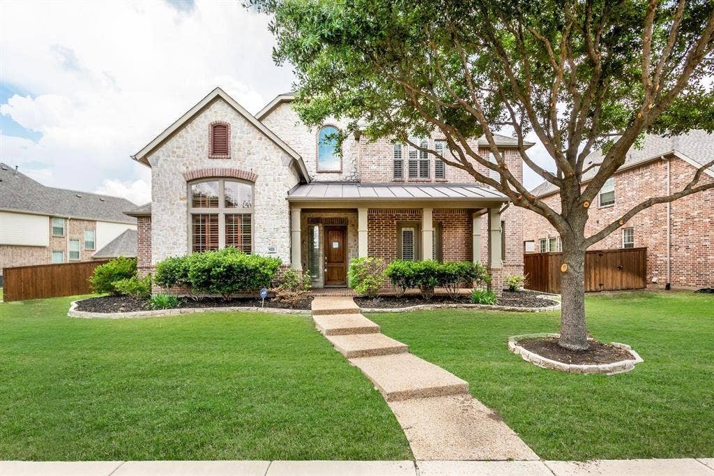 921 Pine Burst Drive, Allen, TX 75013 Stone and brick facade residence featuring a covered front porch, mature front yard tree, arched window details, and a concrete walkway