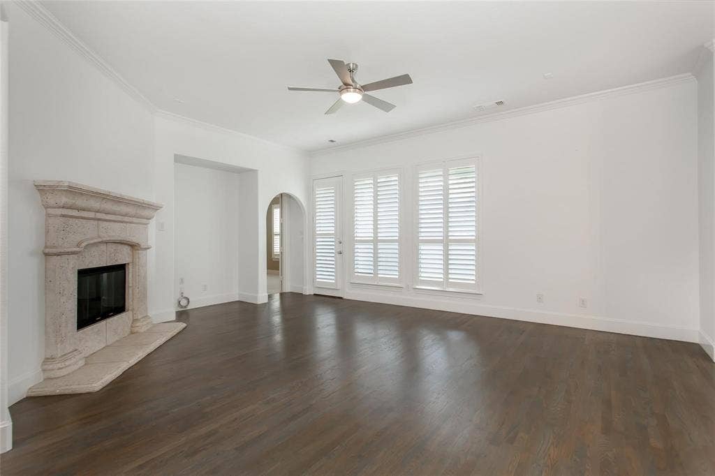 921 Pine Burst Drive, Allen, TX 75013 Spacious room featuring dark wood-finish flooring, a stone fireplace with a hearth, multiple windows with white plantation shutters, an arched doorway, and a ceiling fan