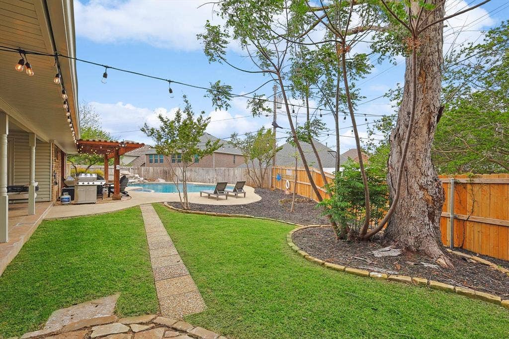 1208 Mayleaf Drive, Irving, TX 75060 Expansive backyard featuring a stone pathway, mature trees, and a wooden pergola covering a built-in outdoor kitchen