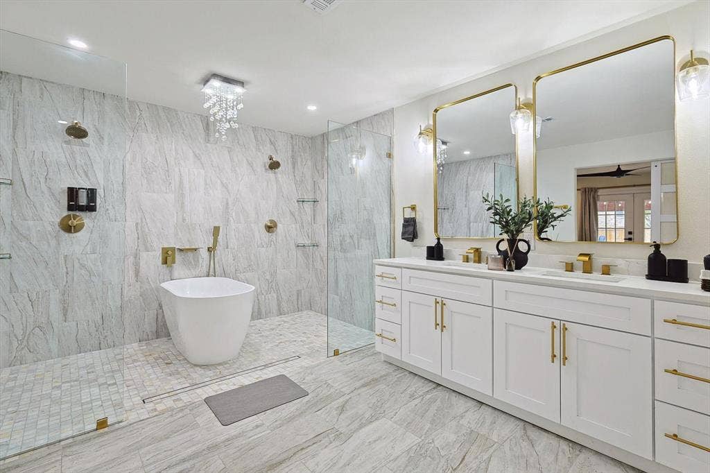 1208 Mayleaf Drive, Irving, TX 75060 Luxurious bathroom featuring a freestanding soaking tub, a spacious walk-in shower with multiple showerheads, a dual vanity with white cabinetry and gold-toned hardware, and large wall-mounted mirrors with sconce lighting