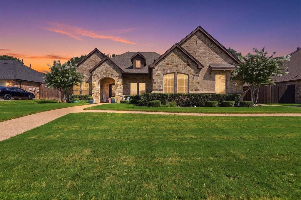 Stone facade residence featuring multiple gables, an arched entry, a concrete pathway, a manicured lawn, and established landscaping
