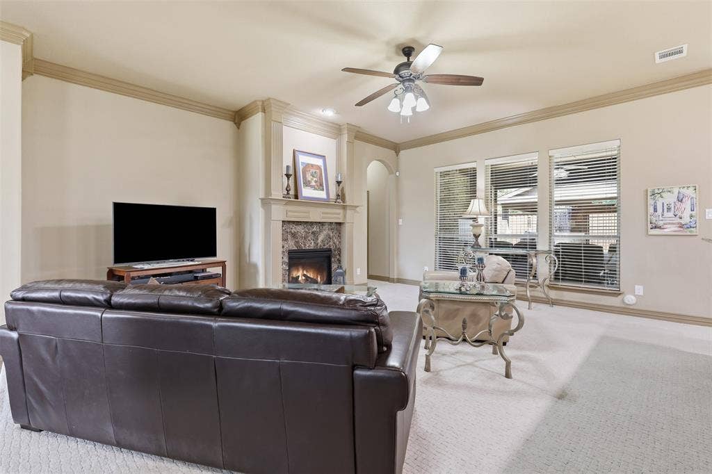 Living area featuring a stone-faced fireplace with a wood mantle, triple windows with horizontal blinds, a ceiling fan, and crown molding
