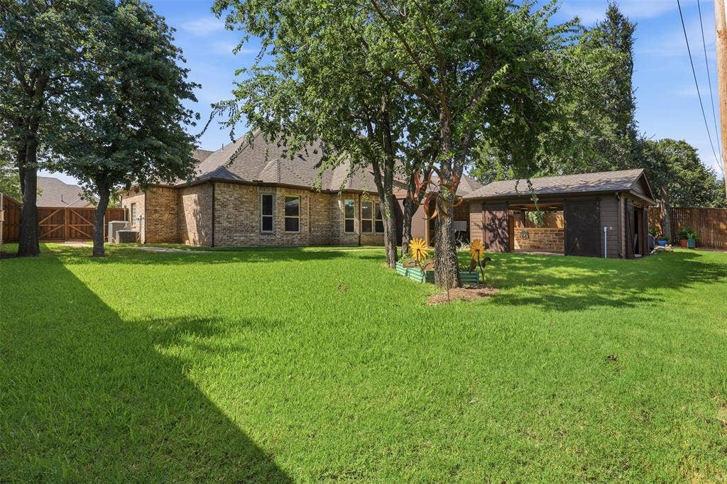 Expansive rear yard featuring a brick-clad residence, mature trees, a detached outbuilding with an open-air covered section, and a wood privacy fence