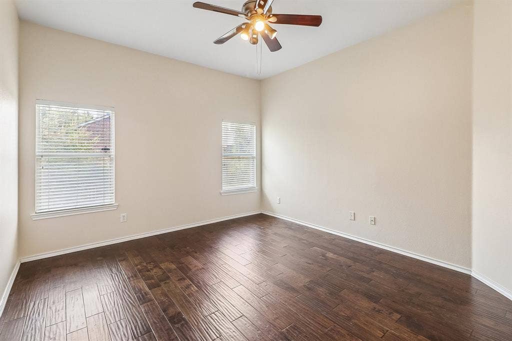 Spacious room featuring rich wood-finish flooring, two windows with blinds, a ceiling fan with integrated lighting, and neutral wall tones