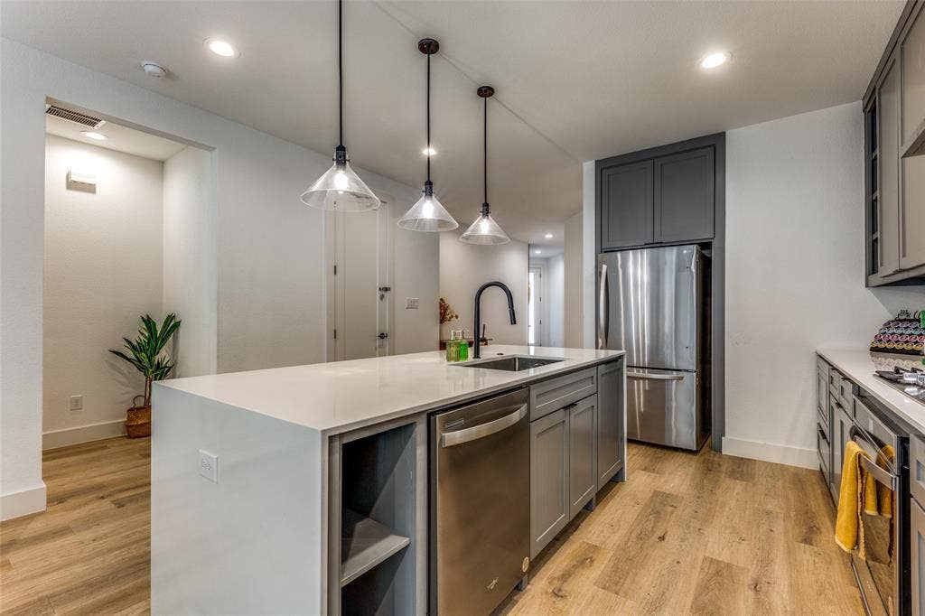 1116 Muncie Avenue, Dallas, TX 75212 Modern kitchen featuring an island with a light-toned countertop, stainless steel appliances, dark gray cabinetry, three pendant lights, and wood-finish flooring
