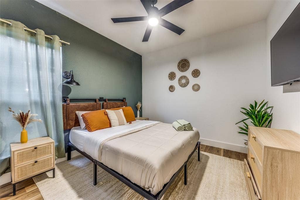 1116 Muncie Avenue, Dallas, TX 75212 Bedroom featuring an accent wall, window with curtains, wood-finish flooring, ceiling fan with integrated lighting, and a wall-mounted television
