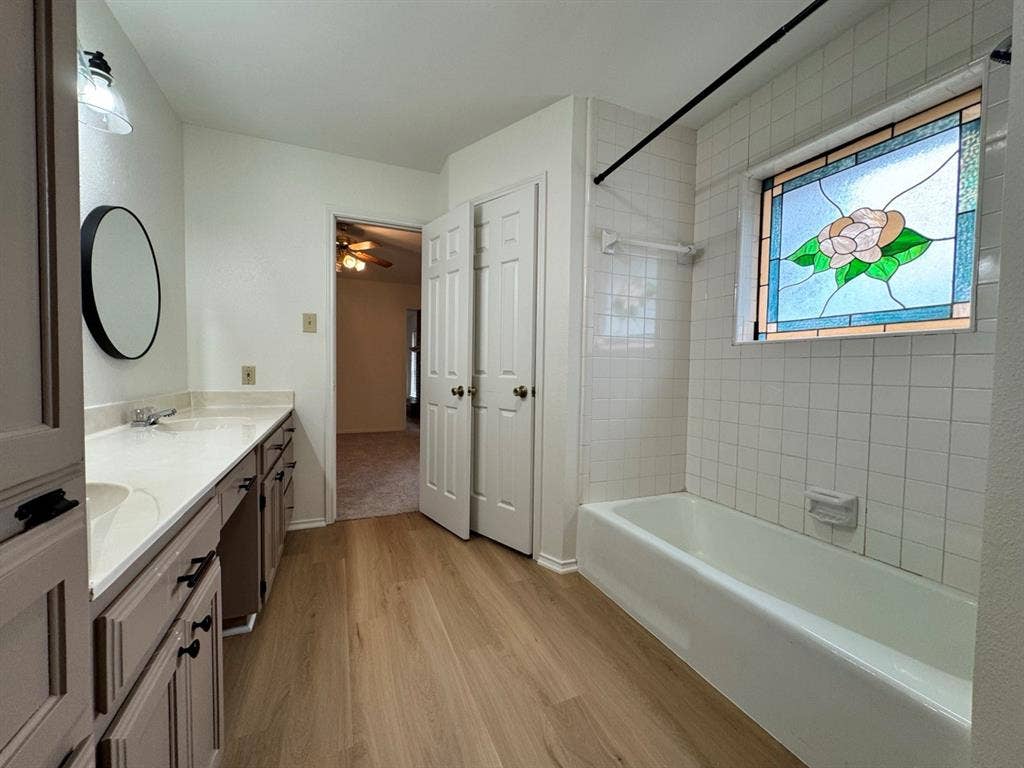 2812 Howard Court, Denton, TX 76209 Bathroom with wood-finish flooring, a double vanity with light countertops, and a white tiled tub surround featuring a decorative stained-glass window