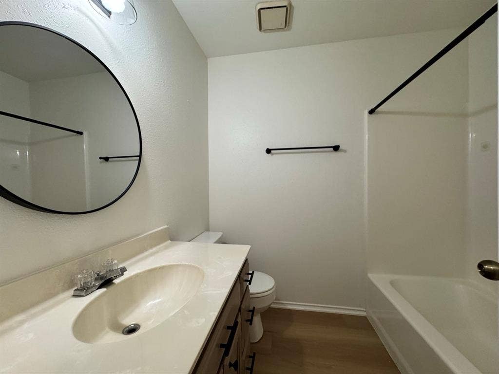 2812 Howard Court, Denton, TX 76209 Bathroom featuring a wood-finish vanity with an integrated sink, a large circular mirror with a black frame, a white bathtub with a shower surround, and light wood-finish flooring