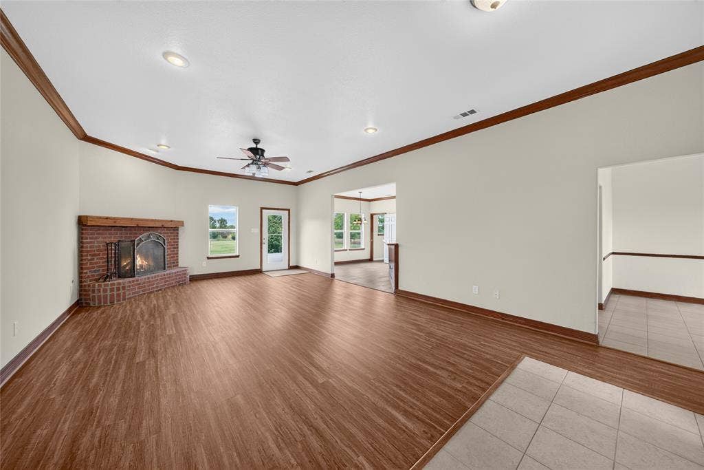 18723 Sims Lane, Nevada, TX 75173 Spacious living area featuring wood-finish flooring, a brick fireplace with a wood mantle, white walls, and dark wood crown molding