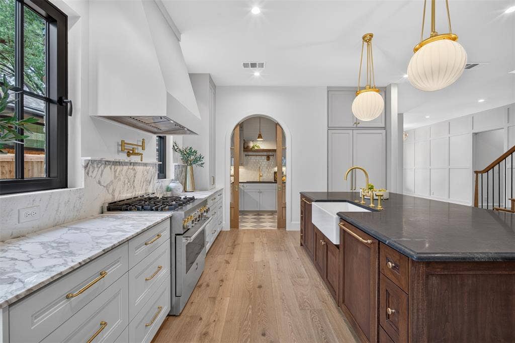 6817 Blessing Drive, Dallas, TX 75214 Gourmet kitchen featuring a large central island with a farmhouse sink, stainless steel range, light wood-finish flooring, marble-finish countertops and backsplash, and a custom range hood