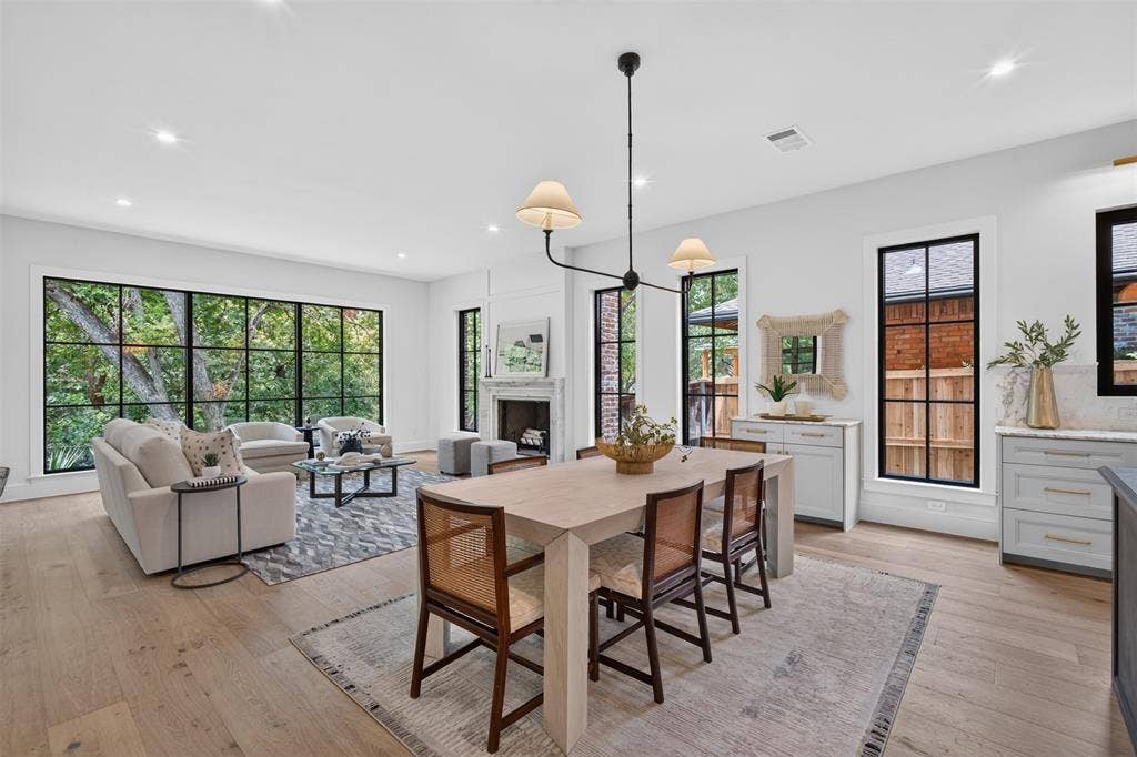 6817 Blessing Drive, Dallas, TX 75214 Open-concept living area featuring light wood-finish flooring, a contemporary chandelier, a white stone fireplace surround, and extensive black-framed windows