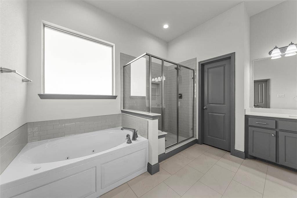Bathroom featuring a corner soaking tub with dark-finish fixtures, a separate glass-enclosed shower, a vanity with dual sinks, and light tile flooring