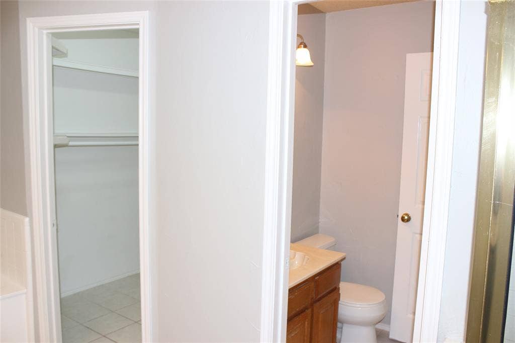 285 Alex Drive, Coppell, TX 75019 Interior view of a closet with white shelving and hanging rod, alongside a bathroom featuring a vanity with a light-toned countertop, a toilet, and a brass-toned wall sconce