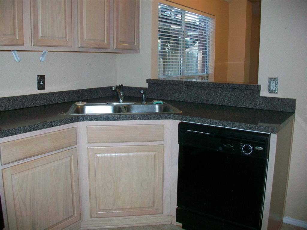 4708 Topaz Lane, Granbury, TX 76049 Kitchen workstation featuring a double-basin stainless steel sink, dark speckled countertop, light wood cabinetry, and a built-in black dishwasher
