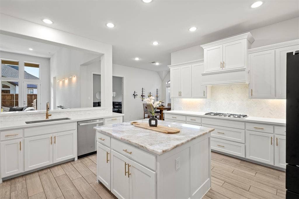 2837 Midlake Drive, Midlothian, TX 76065 Contemporary kitchen featuring a large central island with a stone countertop, white cabinetry with gold-tone hardware, wood-finish flooring, a stainless steel dishwasher, and a gas range with an integrated hood