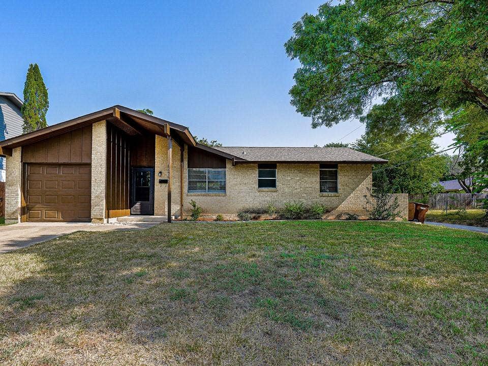 1205 Northridge Drive, Plano, TX 75075 Brick ranch-style home featuring a single-car attached garage with wood-finish door, manicured front lawn, and mature landscaping including established trees and evergreen plantings