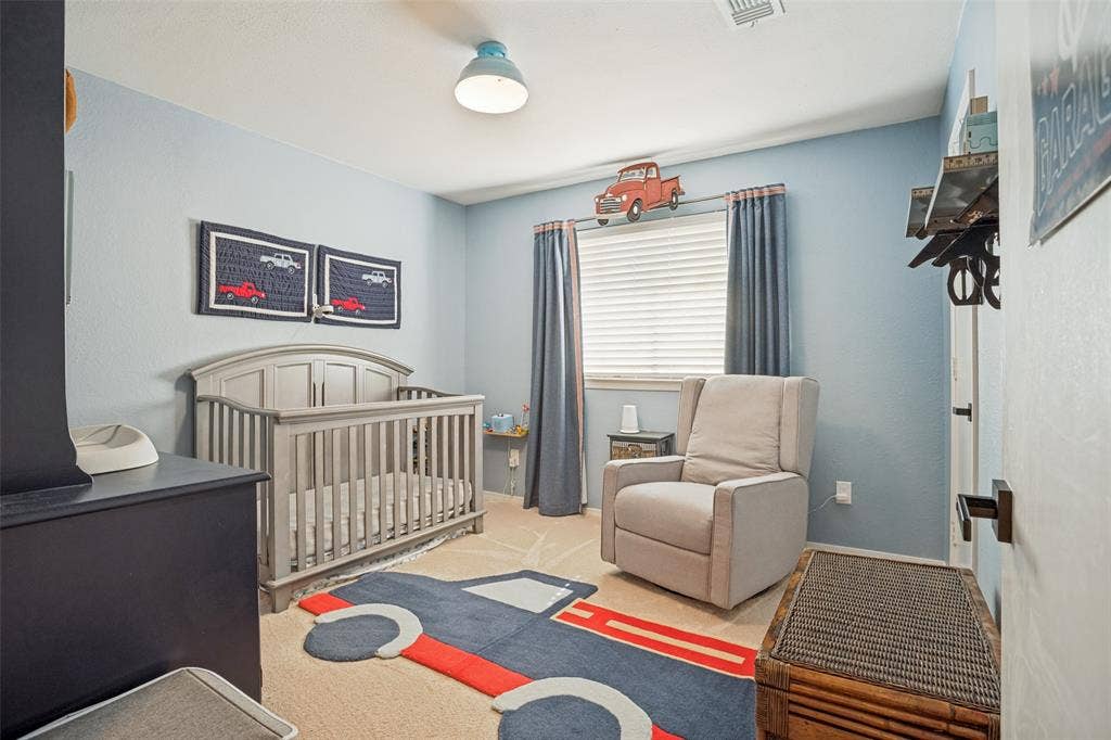 8201 Christie Drive, Frisco, TX 75033 Bright nursery bedroom featuring soft blue walls, natural light from a large window with gray curtains, wood-finish flooring with coordinating area rug, wooden crib, upholstered glider chair, and ceiling-mounted fixture