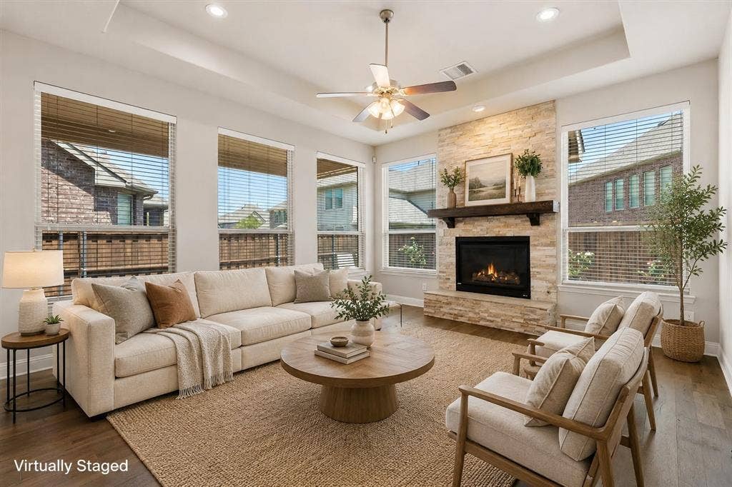 4109 Rainey Street, McKinney, TX 75070 Spacious living area featuring a stone-faced fireplace with a wood mantel, multiple large windows, a ceiling fan, and wood-finish flooring