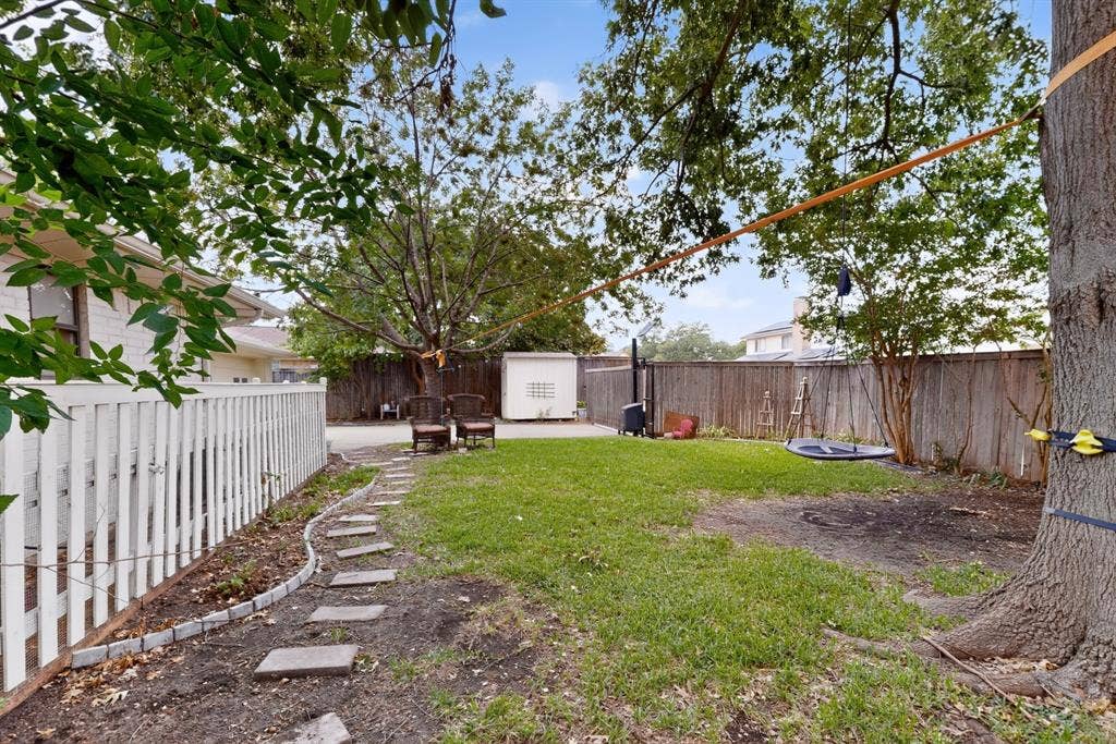 2003 J J Pearce Drive, Richardson, TX 75081 Expansive backyard featuring a lush lawn, mature shade trees, a white picket fence, a stepping stone pathway, and a storage shed