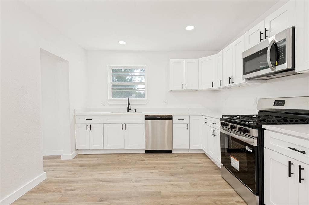 Modern kitchen featuring white shaker cabinetry with matte black hardware, light wood-finish flooring, stainless steel appliances, and a black faucet