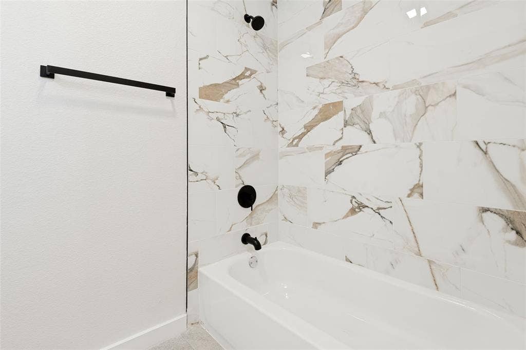 White bathtub with matte black fixtures, surrounded by large format porcelain wall tile featuring white, brown, and gray veining