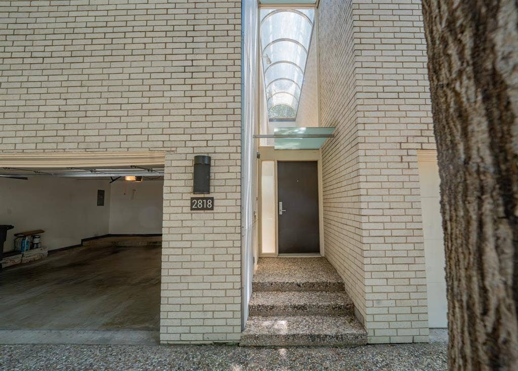 2818 Welborn Drive, #9, Dallas, TX 75219 Light-colored brick exterior featuring an attached garage, modern front entry with an overhead glass canopy, and an arched skylight above the entry steps