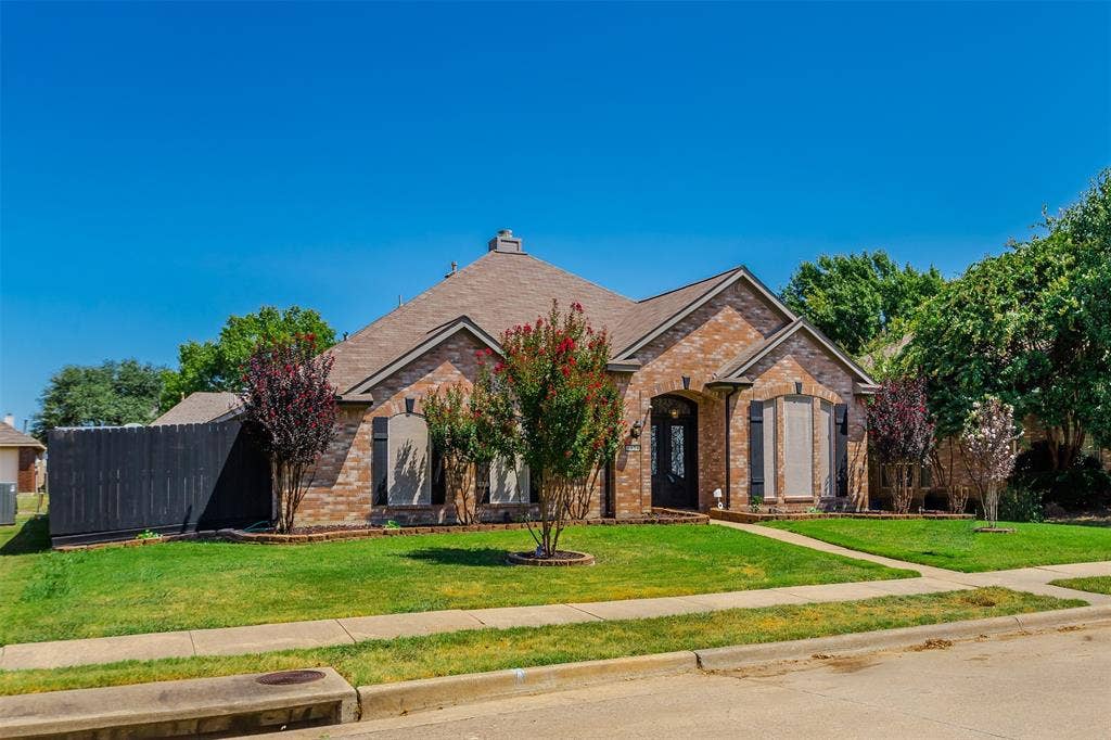 11404 Amber Valley Drive, Frisco, TX 75035 Brick Colonial home showcasing a manicured front lawn, mature landscaping with flowering shrubs and trees, a gabled roofline with architectural detailing, shuttered windows, and a curved driveway approach