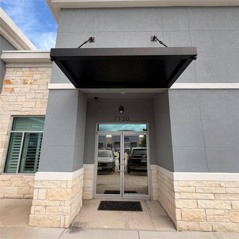 945 Stockton Drive, #7120, Allen, TX 75013 Modern commercial entrance featuring a black overhead awning, prominent building numbers, a light gray stucco facade, and a stone veneer base