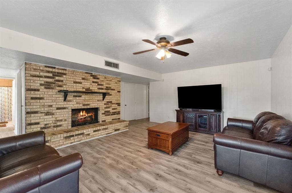 Brick-faced fireplace with a hearth, wood-finish flooring, and a ceiling fan with integrated lighting
