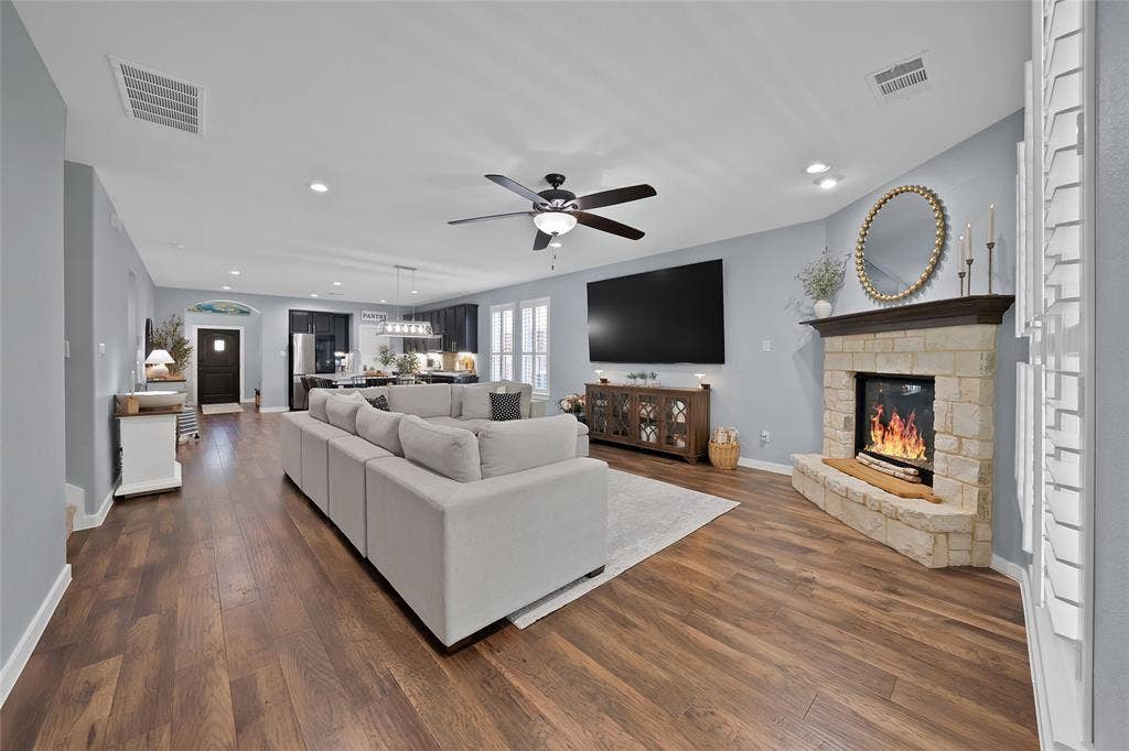 Spacious open-concept living area featuring wood-finish flooring, a corner stone-faced fireplace with wood mantel, recessed lighting, and a ceiling fan
