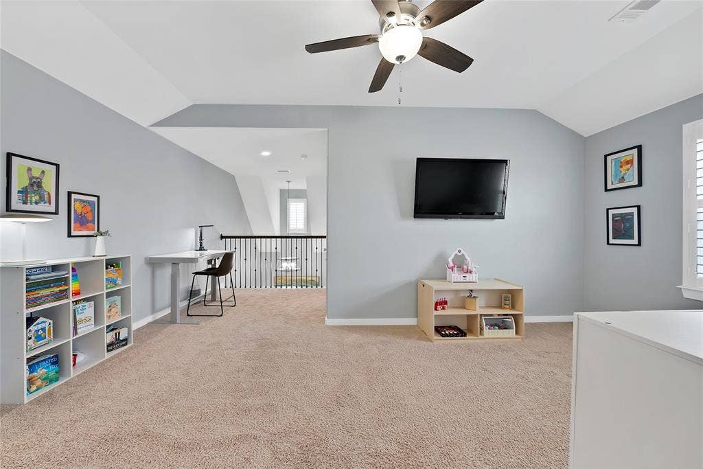 Carpeted loft area featuring a vaulted ceiling with a ceiling fan, light gray walls, an open railing overlooking a lower level, and a window with plantation shutters