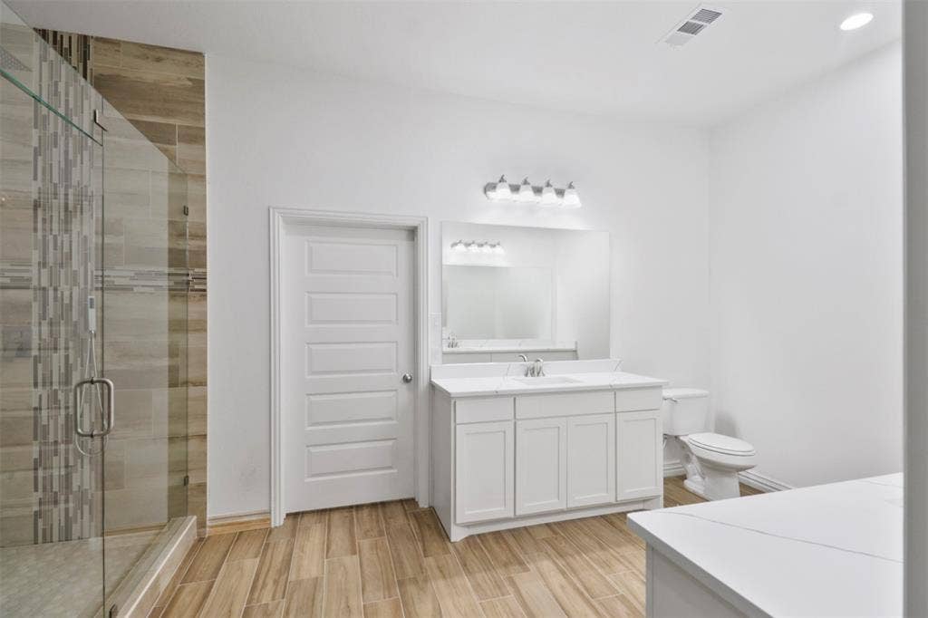 4112 Balch Drive, Dallas, TX 75216 Spa-inspired primary bathroom featuring a frameless glass-enclosed shower with mixed-material tile surround, dual-sink vanity with white cabinetry and integrated lighting, wood-finish flooring, and a freestanding soaking tub