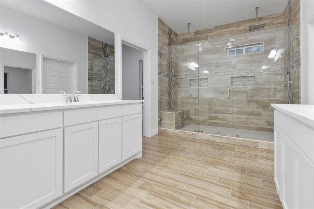 4112 Balch Drive, Dallas, TX 75216 Spa-inspired primary bathroom featuring dual vanities with white cabinetry, a frameless glass-enclosed walk-in shower with tile surround and rainfall showerhead, wood-finish flooring throughout, and abundant natural lighting from transom windows