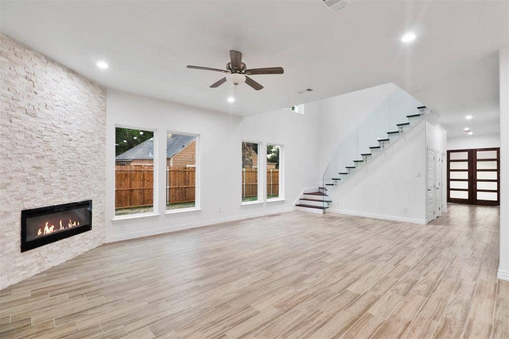 4112 Balch Drive, Dallas, TX 75216 Spacious living area featuring wood-finish flooring, a linear fireplace with a stacked stone surround, multiple windows, a ceiling fan, and a floating staircase with glass railings