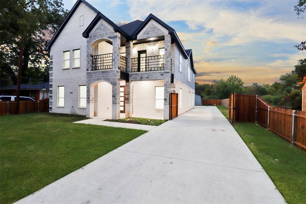 4112 Balch Drive, Dallas, TX 75216 Contemporary exterior featuring white brick and stone accents, a dual second-story balcony with iron railings, a stone archway entryway, a concrete driveway, and a wooden privacy fence