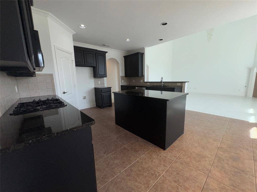 1913 Lawnview Drive, McKinney, TX 75072 Modern kitchen featuring a substantial black island with waterfall edge, gas cooktop with stainless steel range hood, dark cabinetry, subway tile backsplash, and wood-finish flooring throughout the open layout