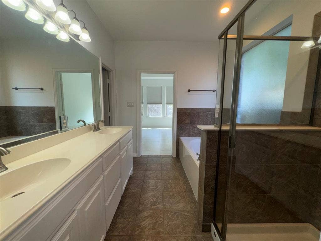 1913 Lawnview Drive, McKinney, TX 75072 Bathroom featuring a dual vanity with white cabinetry and integrated sinks, a large wall-mounted mirror, a glass-enclosed shower, a soaking tub, and floor-to-ceiling tile accents