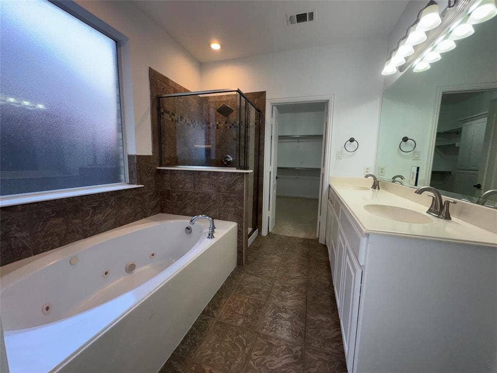 1913 Lawnview Drive, McKinney, TX 75072 Spa-inspired master bathroom featuring a soaking tub with glass enclosure and tile surround, dual vanities with white cabinetry and undermount sinks, large framed mirror with integrated lighting, and dark tile flooring throughout