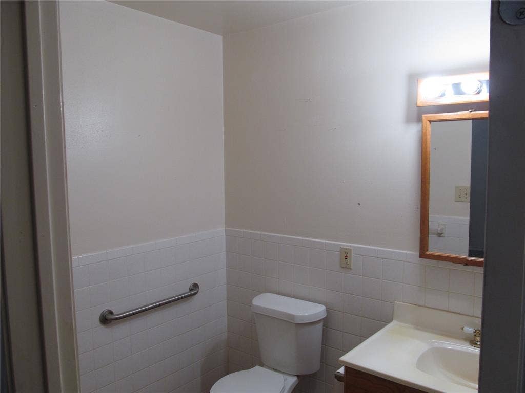740 Emberwood Drive, Dallas, TX 75232 White tile wainscoting, built-in grab bar, wood-finish vanity with single sink, and wall-mounted mirror with integrated lighting