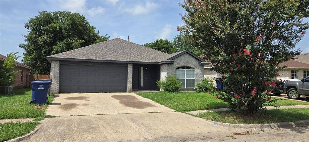 4809 Orchard Ridge Drive, Garland, TX 75043 Gray brick exterior with a dark gray garage door, a gray shingle roof, a front lawn, and a mature tree with pink blooms