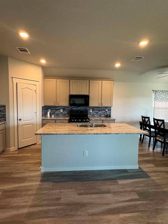 Fort Worth, TX 76123 Open-concept kitchen featuring an island with an undermount sink, overhead recessed lighting, wood-finish flooring, a patterned tile backsplash, and light-toned cabinetry