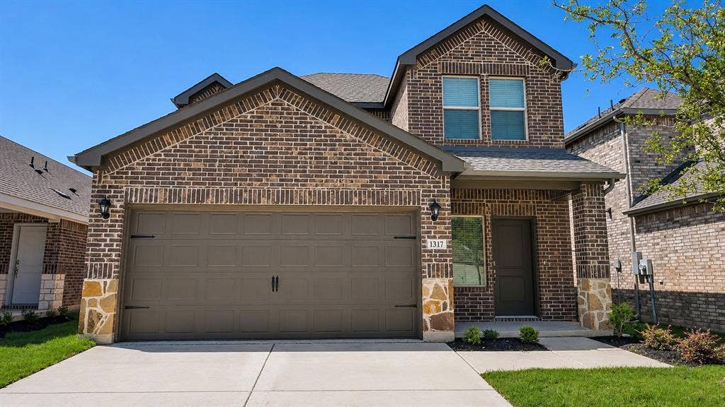 1317 crookpost Drive, Princeton, TX 75407 Stunning two-story brick home featuring a three-car garage with dark panel doors, arched gable roofline with decorative brick detailing, and a professionally finished driveway