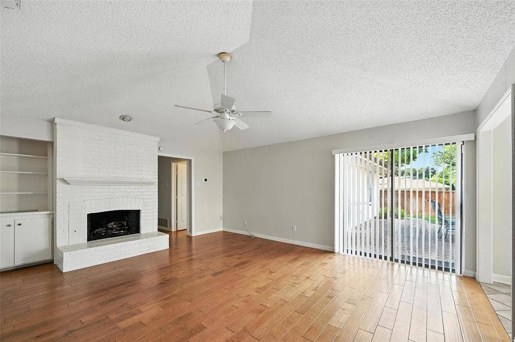 Living area featuring wood-finish flooring, a white painted brick fireplace with hearth, built-in shelving, a ceiling fan, and sliding glass doors with vertical blinds