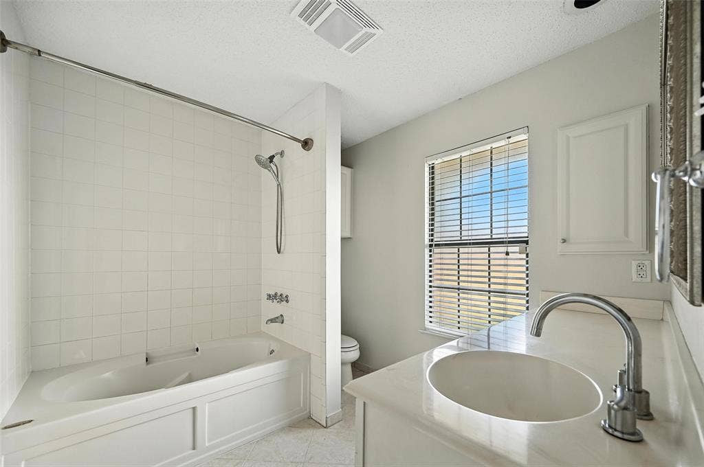 Bathroom featuring a white tiled tub and shower combination with a handheld showerhead, a single vanity with an integrated sink and chrome faucet, and a window with horizontal blinds