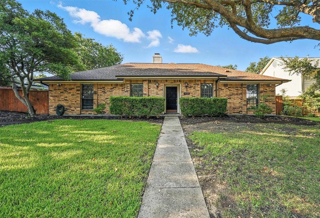 Charming brick ranch home featuring a manicured front lawn, mature shade trees, and a welcoming center entry with black-trimmed windows and doors