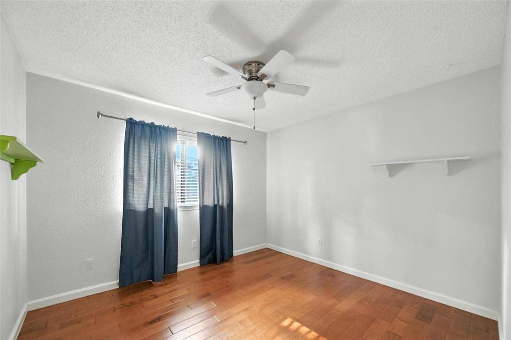 Spacious bedroom showcasing warm wood-finish flooring, ceiling fan with integrated lighting, and dual windows dressed in layered drapery that provide natural light and privacy control