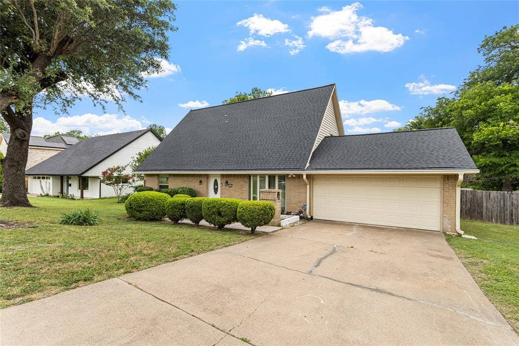 6930 Trailcrest Drive, Dallas, TX 75232 Brick facade home featuring a two-car garage, dark shingle roof, mature front yard tree, manicured shrubbery, and a concrete driveway