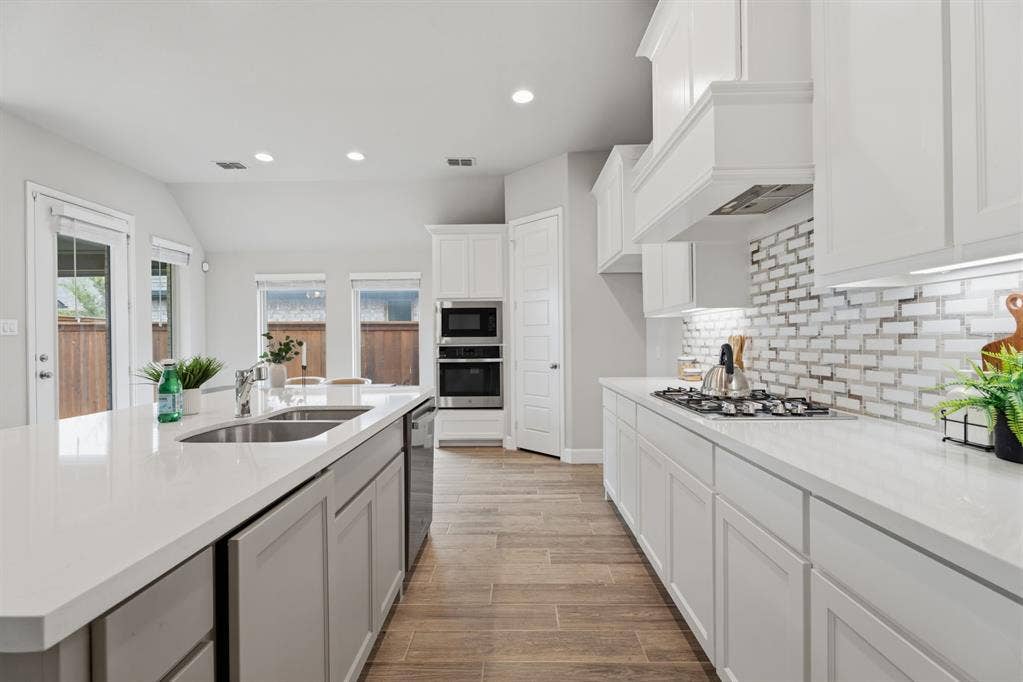 Bright kitchen featuring an expansive island with a double sink, white cabinetry, integrated stainless steel appliances, a gas cooktop, and a mosaic tile backsplash