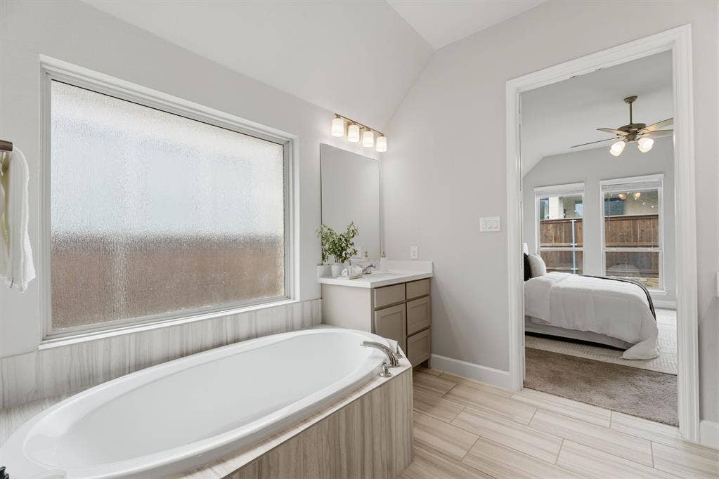 Freestanding soaking tub with tiled surround, large privacy window, dual vanity, and wood-finish flooring