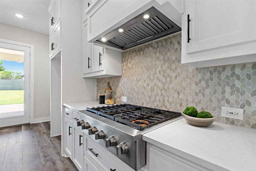 1821 Stonewall Road, Argyle, TX 76226 Stainless steel gas range with coordinating ventilation hood, white cabinetry with matte black hardware, mosaic tile backsplash, light-toned countertops, and wood-finish flooring